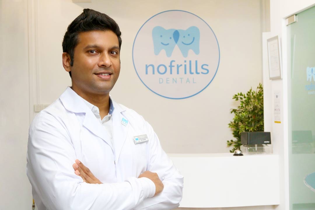 dr-daneshkumar-at-nofrillsdental-marina-square dentist standing in front of signage of nofrills dental